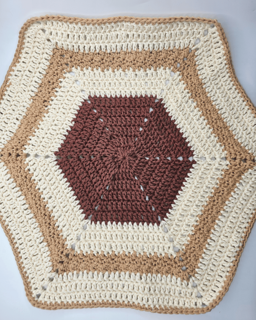 The blanket part of the monkey lovey crochet pattern completed is laid flat on a surface. It is hexagon in shape with a mix of double crochet stitches, lemon peel stitch, and a single crochet border. It is made from three colours: dark brown in the centre, cream, then light brown, cream, then a light brown border.